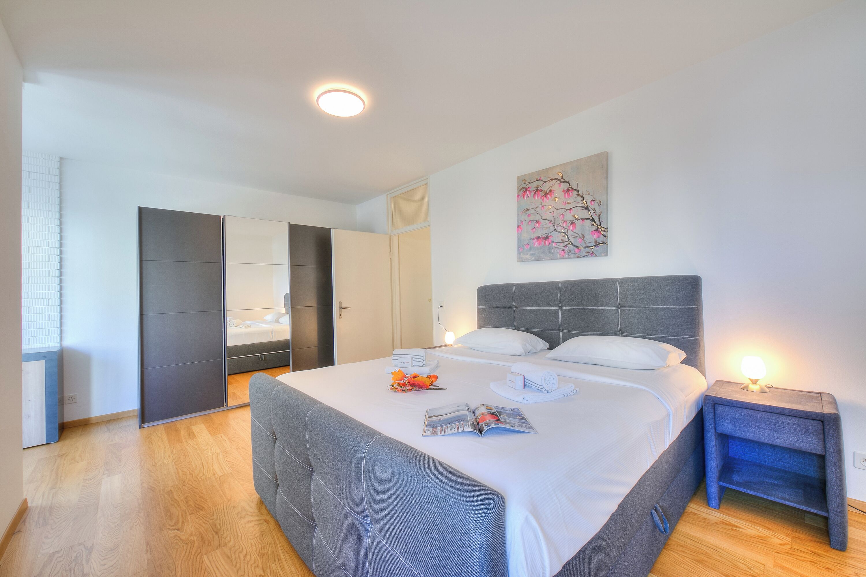 A modern bedroom with a large bed, a bedside table with a lamp, a wall-mounted artwork, and a sliding door leading to another room.