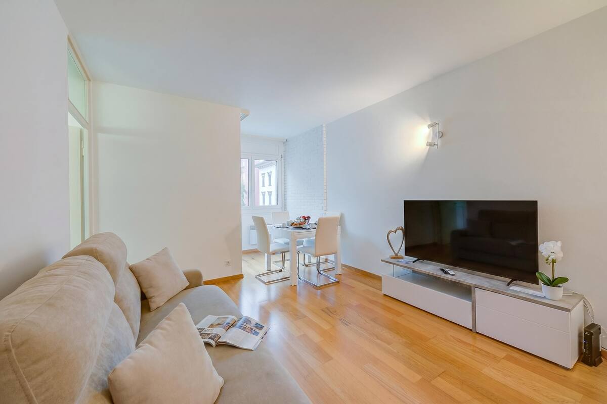 A modern living room with a sofa, a TV, and a dining area in the background.