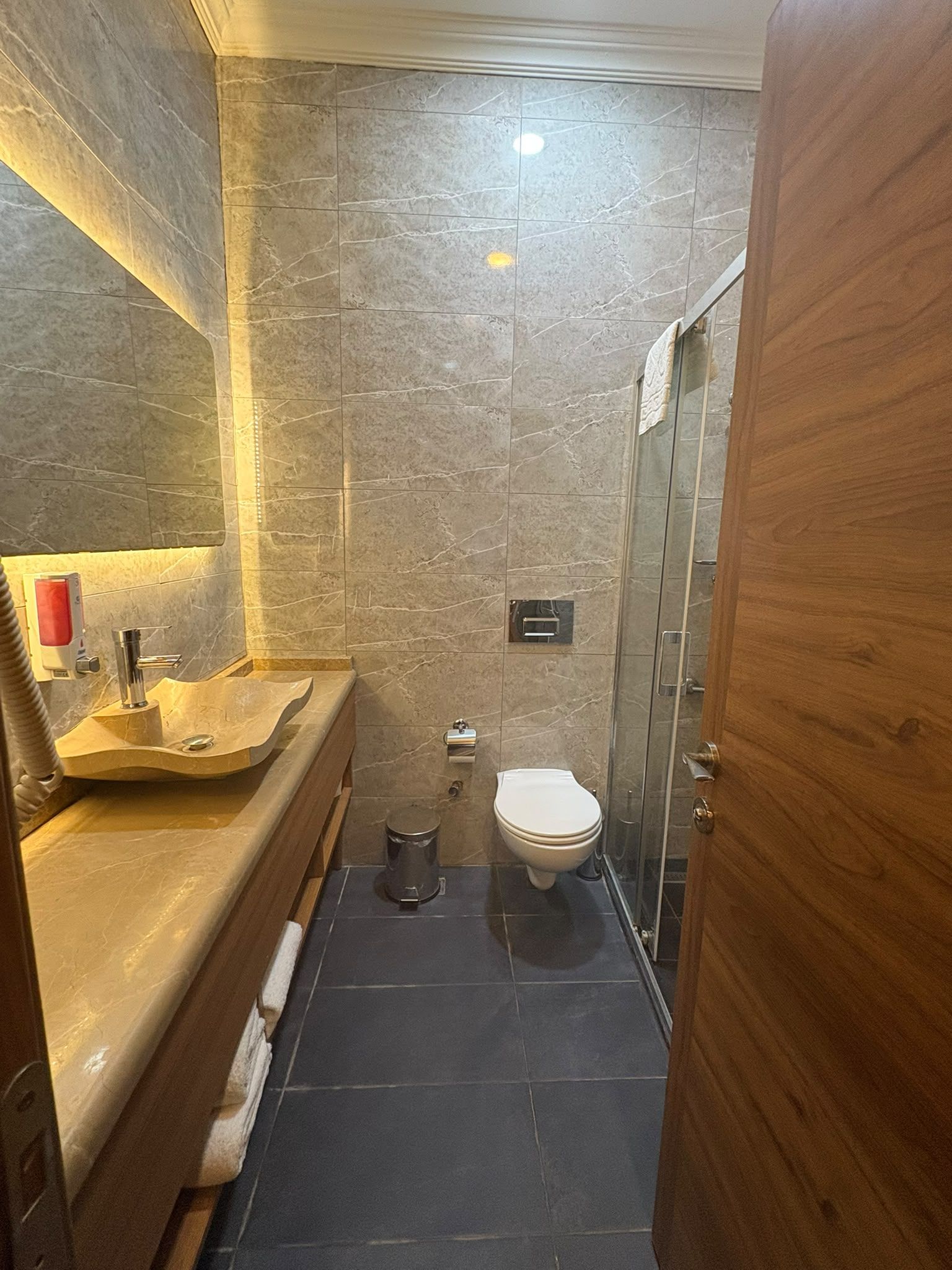 Une salle de bain avec un mur en marbre, un meuble-lavabo en bois, des toilettes et un espace douche.