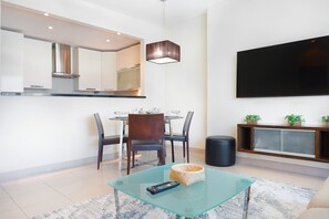 Un salon moderne avec une table basse en verre, un coin repas avec des chaises, un téléviseur fixé au mur et un espace cuisine avec des placards et une cuisinière.