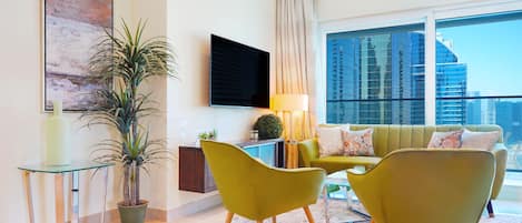 Una sala moderna con un sofá, dos sillones, una mesa de centro de vidrio, un televisor, una lámpara, una planta en maceta y un ventanal con vistas a la ciudad.