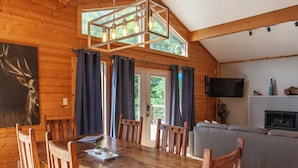 A wooden dining room with a large table and chairs, a chandelier, and a kitchen area with a TV and fireplace. - Stackstone Ridge- Gorgeous View! (Bryson City)