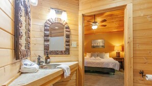 A wooden cabin interior with a bathroom featuring a sink, mirror, and towel rack. A bedroom with a bed, nightstand, and lamp is visible through an open door. - Stackstone Ridge- Gorgeous View! (Bryson City)