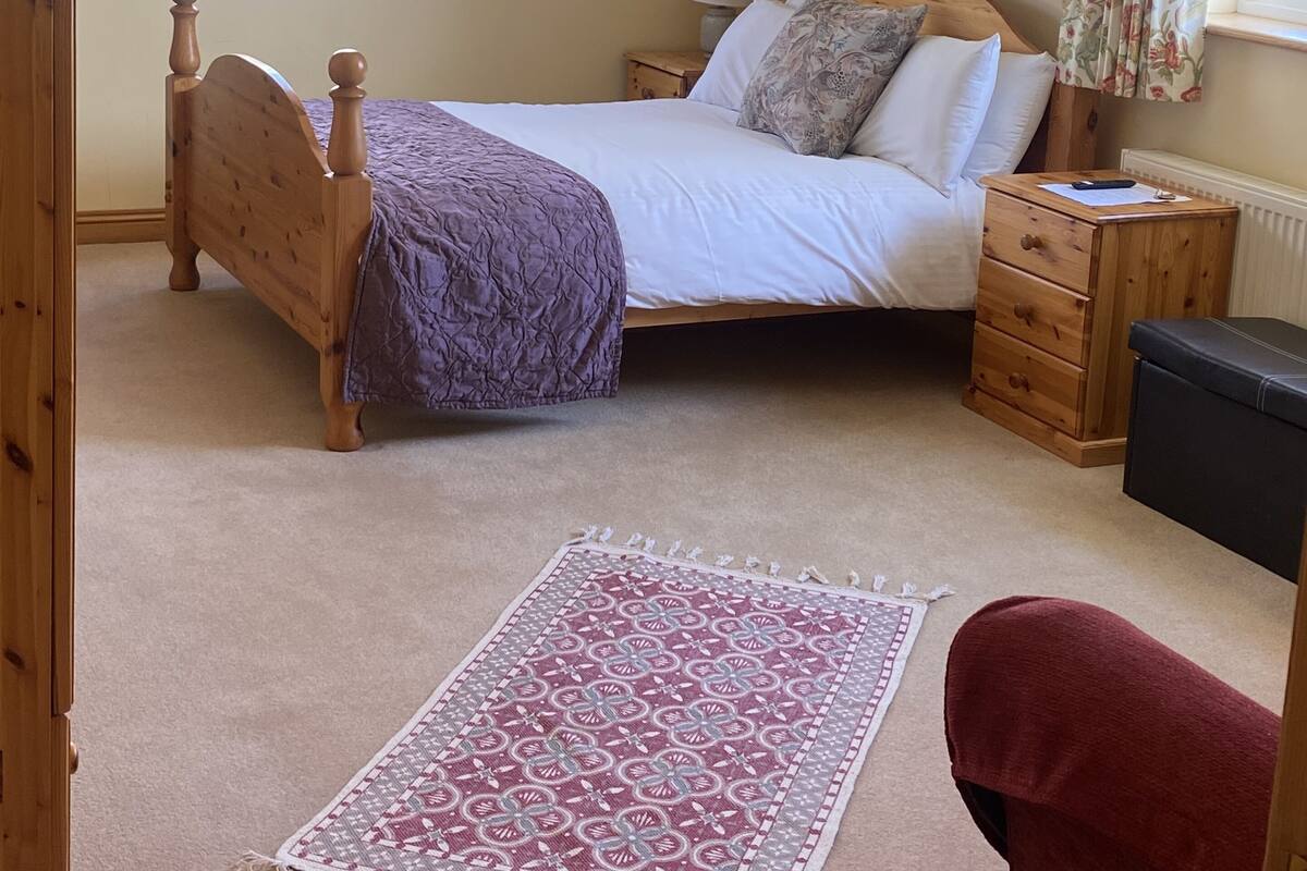 Un dormitorio con techo inclinado, una cama con cabecera de madera, una mesita de noche, un aparador, una ventana con cortinas florales y un tragaluz.