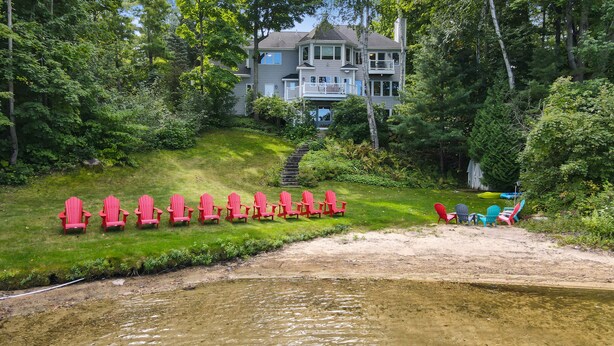 Propiedad frente al lago con una casa, una zona verde con sillas Adirondack rojas y una playa de arena con sillas coloridas.
