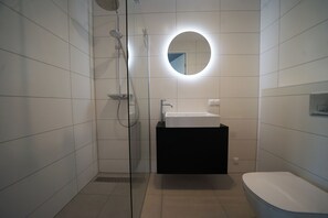 Et moderne badeværelse med et glasbadeværelsesafskærmning, et rundt spejl, en hvid vask og et toilet.