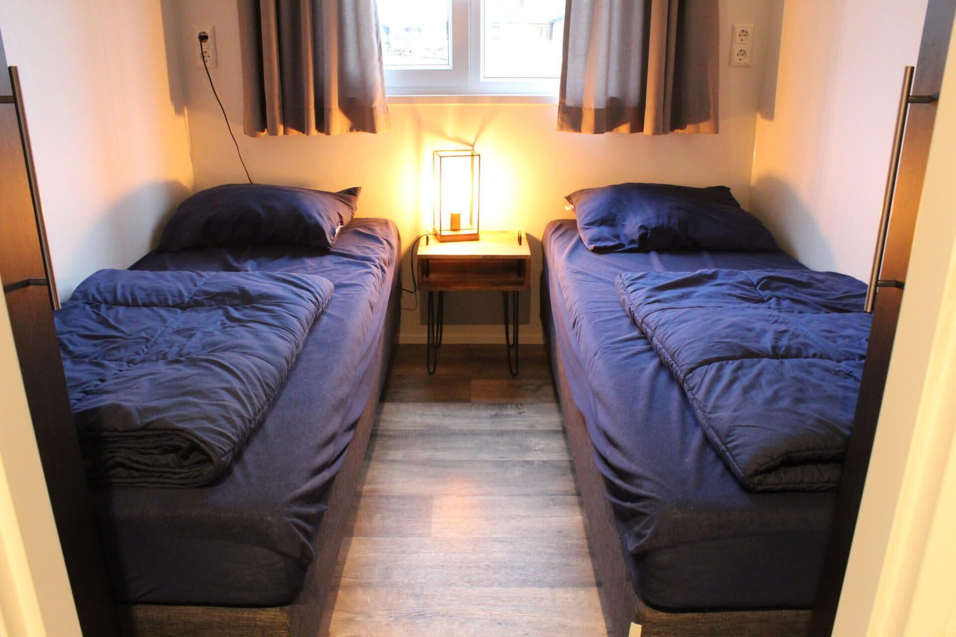 A narrow room with two beds, a small table with a lamp, and a window with curtains.