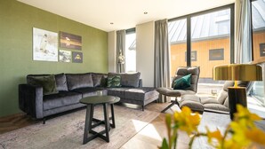 Una sala de estar moderna con un sofá seccional gris, una mesa de centro redonda y un sillón gris. Hay ventanas amplias con cortinas, una pared verde con obras de arte y un suelo de madera.