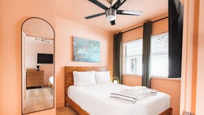 Ein Schlafzimmer mit einem Bett, einem Deckenventilator, einem Gemälde, einem Fenster mit Vorhängen und einer Holzkommode.