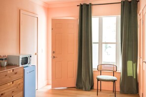 Una habitación con puerta, microondas, refrigerador pequeño, silla y ventana con cortinas.