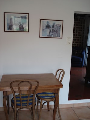 A dining area with a wooden table and chairs, two framed paintings on the wall, and a view of a blue sofa in the background.