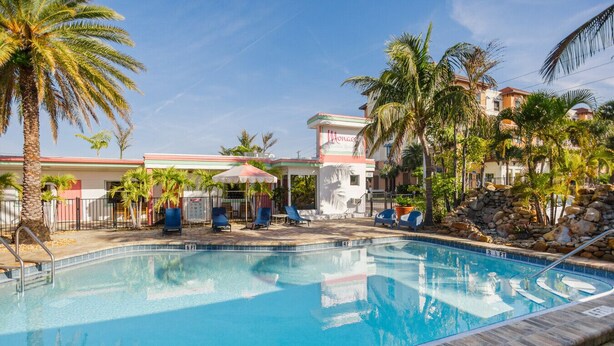 A swimming pool with lounge chairs and umbrellas, surrounded by palm trees and a building with a sign. - 129 · ☀Pool✔ Kitchen✔ Beach✔ Self Checkin✔ Free Parking✔ (Clearwater)