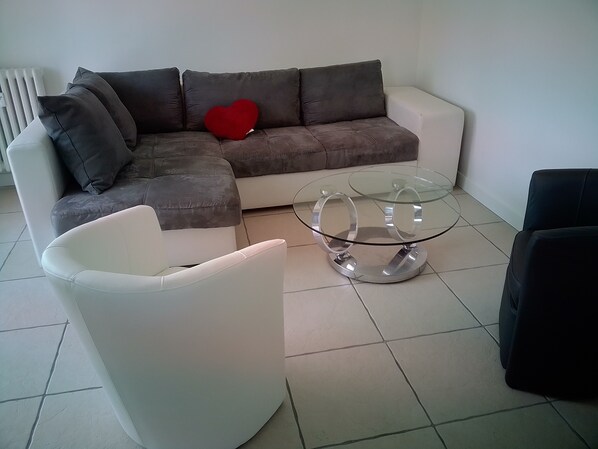 Uma sala de estar com um sofá em forma de L, uma mesa de centro de vidro e uma poltrona branca.