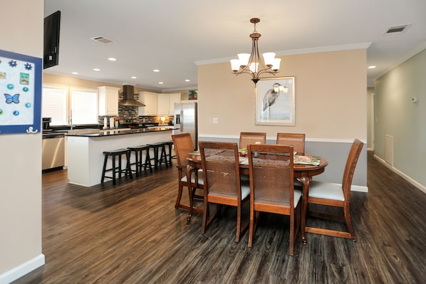 A dining area with a wooden table and chairs, a chandelier, and a kitchen in the background.