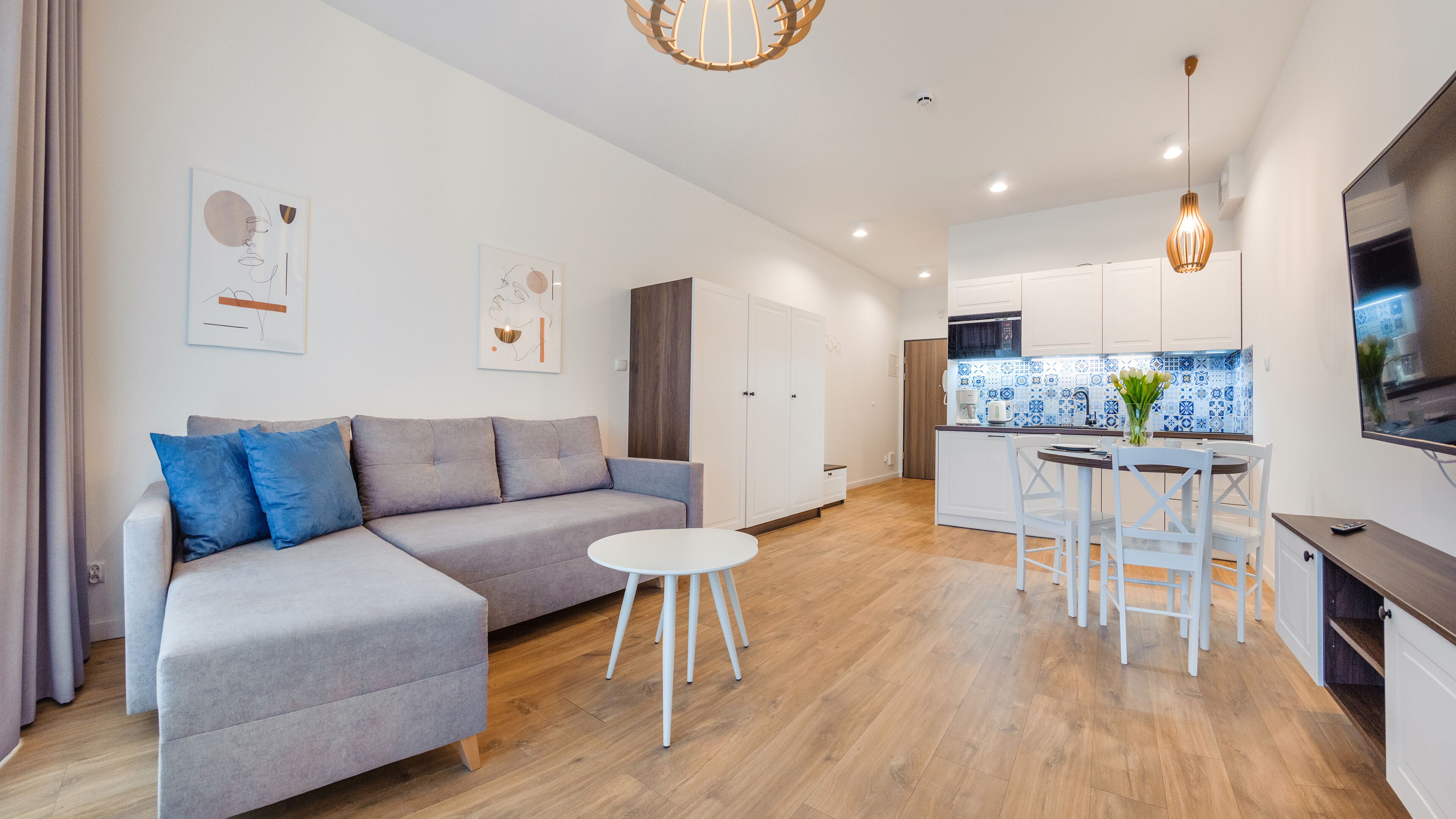 A modern living room with a grey sectional sofa, a small white table, and a kitchen area with white cabinets and blue tiles.