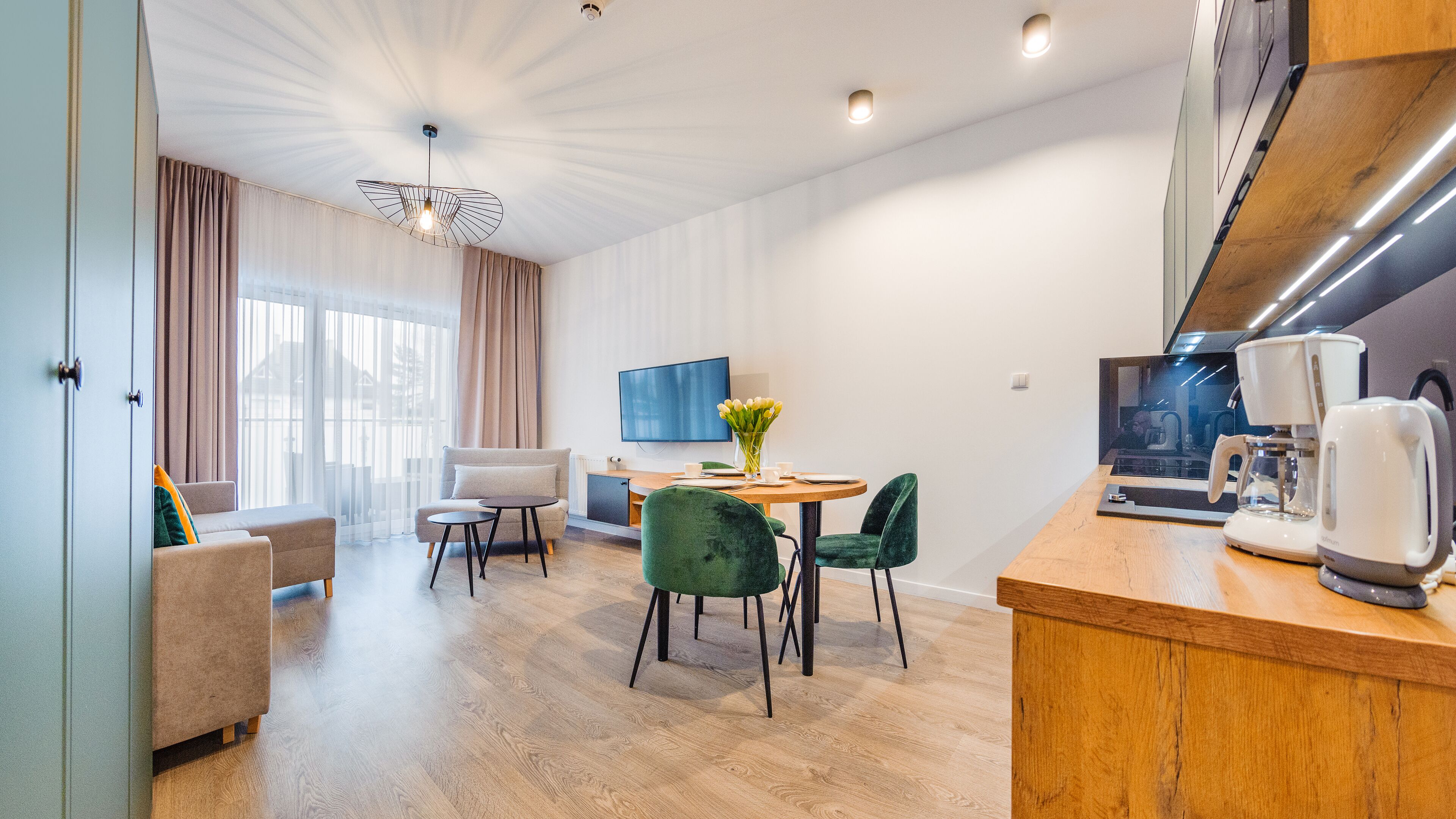 A modern living room with a sofa, a dining table with chairs, a TV, and a kitchen area with a coffee machine.