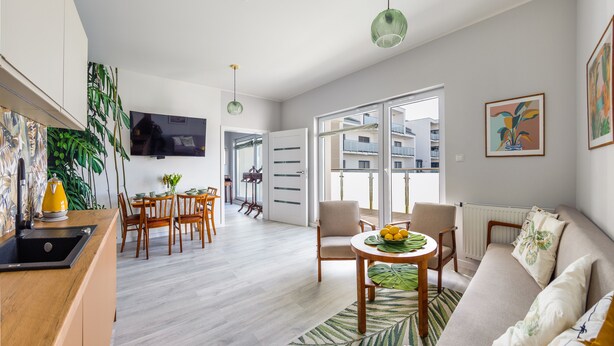 Una sala de estar moderna con sofá, una mesa de comedor con sillas, televisor montado en la pared y un balcón con vista a edificios.