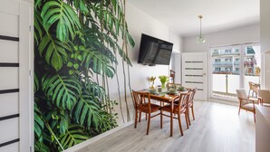 Un comedor con un gran mural en la pared de follaje verde, una mesa de madera con sillas, un televisor de pantalla plana montado en la pared y vista a un balcón a través de una puerta de cristal.