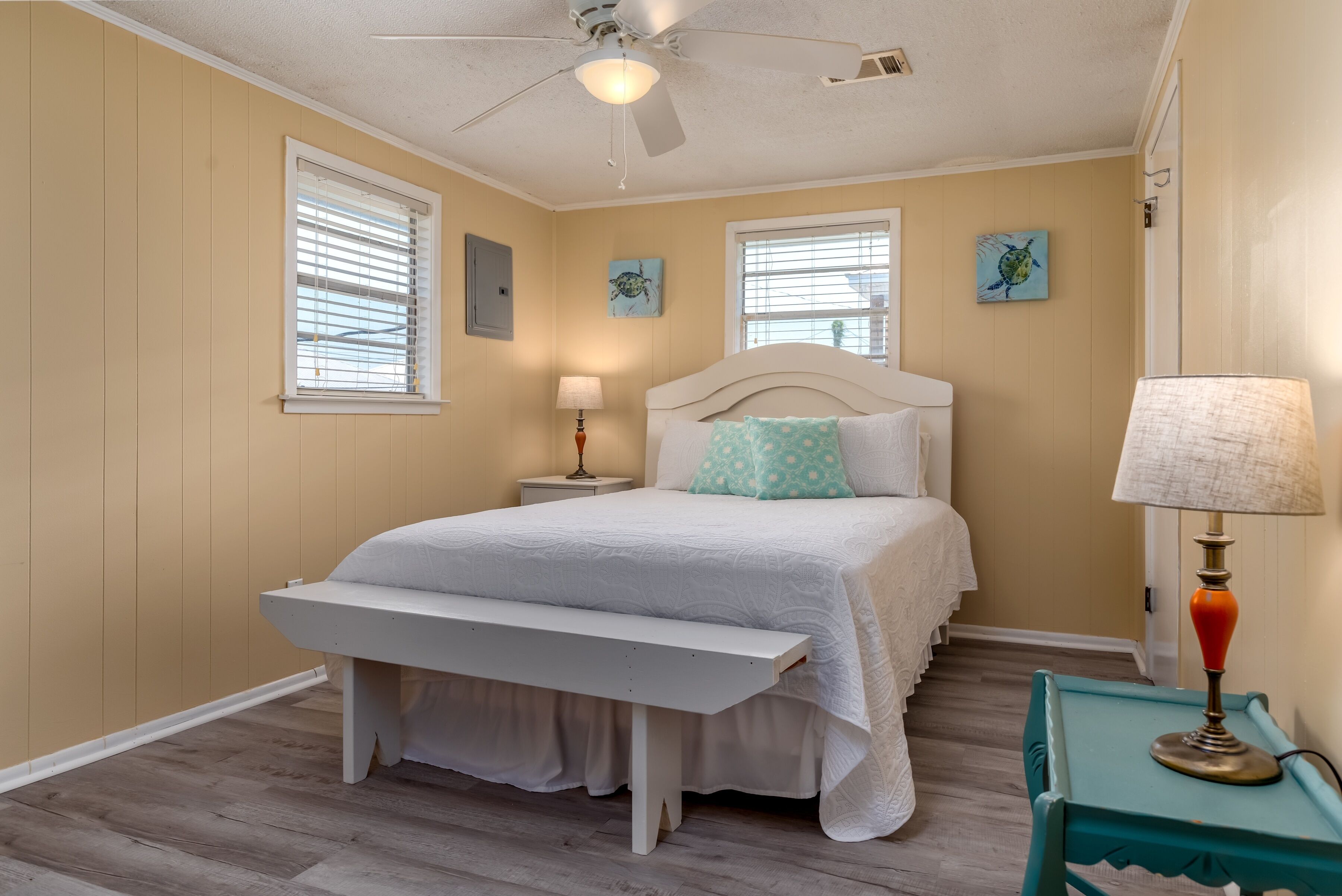 A bedroom with a bed, a bench, a nightstand, a lamp, and two windows.