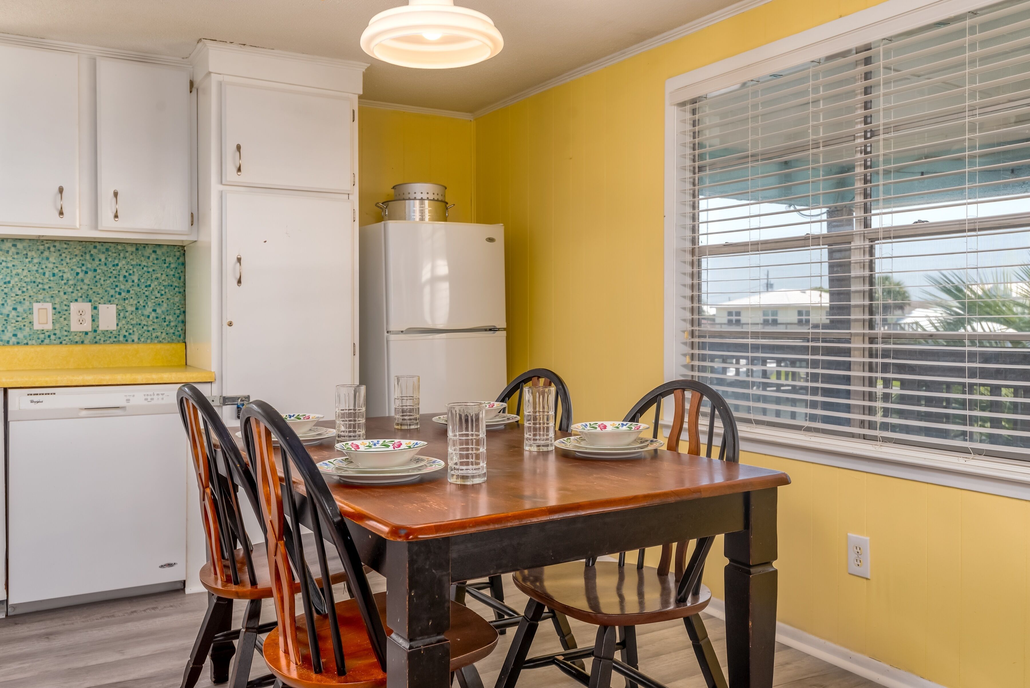 A dining area with a wooden table and chairs, a white refrigerator, and a window with blinds.