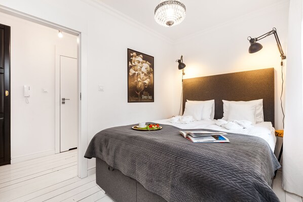 A neatly arranged bedroom with a large bed, a bedside table with a lamp, a framed picture on the wall, and a door leading to another room.