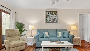 Sala de estar con un sofá azul, un sillón color beige y una mesa de centro blanca. Hay dos lámparas sobre mesas auxiliares, un cuadro en la pared y un ventanal con cortinas.
