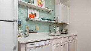 Una cocina con armarios blancos, un lavamanos de acero inoxidable y un lavavajillas. Hay objetos decorativos en la encimera y un cuadro enmarcado sobre el lavamanos.