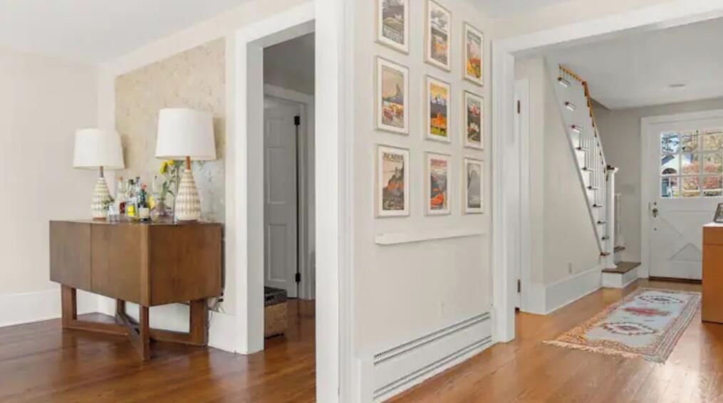 A hallway with a wooden floor, a white wall with framed pictures, a staircase, and a door with a window.