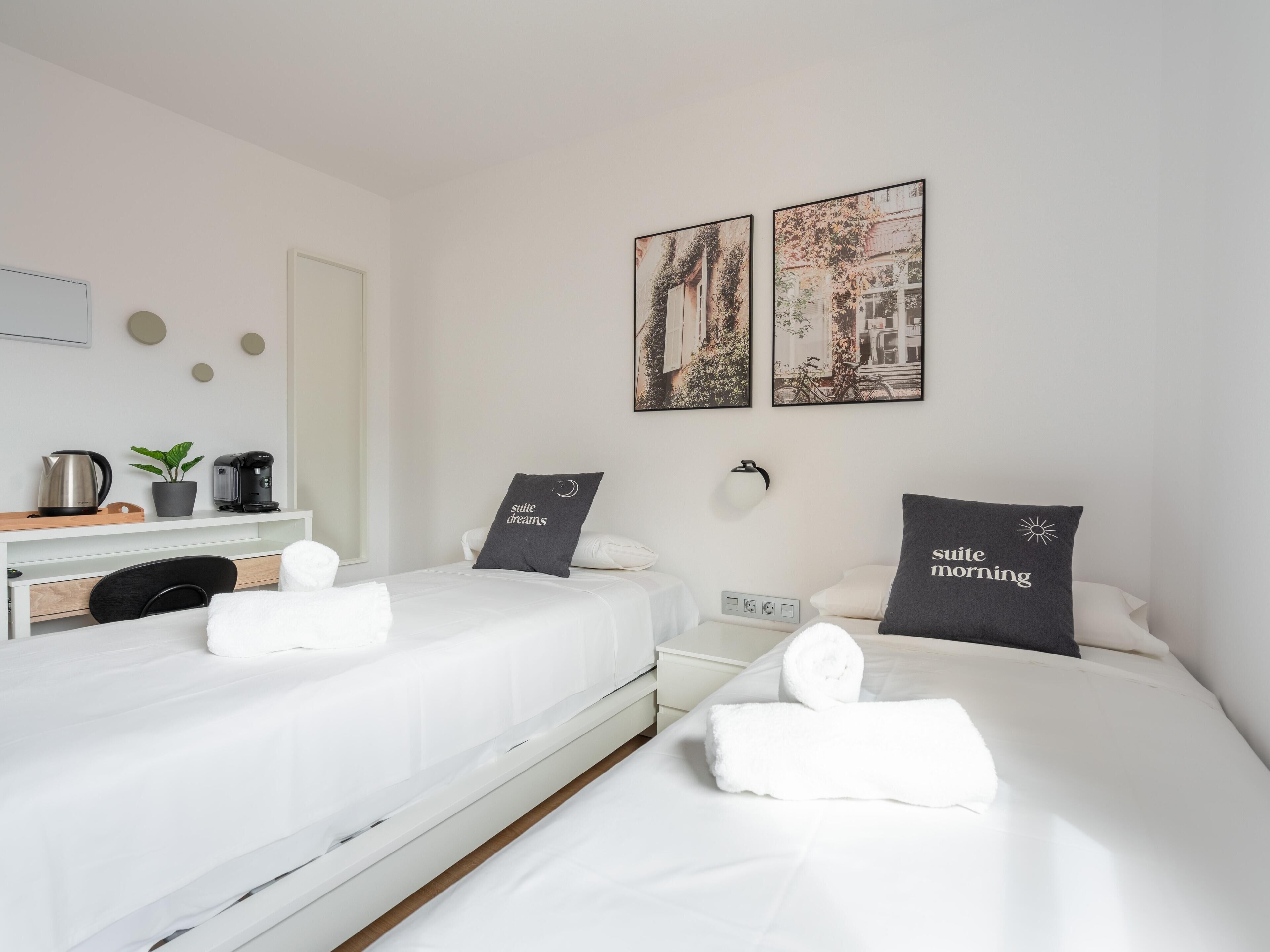 Habitació d'hotel amb dos llits individuals, una taula amb una màquina de cafè, una planta i obra d'art a la paret.