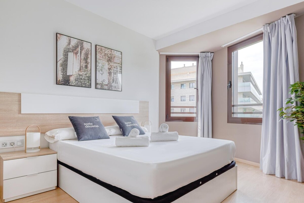 A modern bedroom with a bed, bedside tables, a nightstand, a window with curtains, and artwork on the wall.