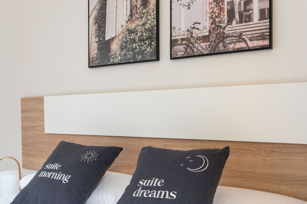 Una cama bien hecha con dos almohadas, cada una con un diseño único. Encima de la cama, dos cuadros enmarcados cuelgan de la pared, mostrando escenas al aire libre.