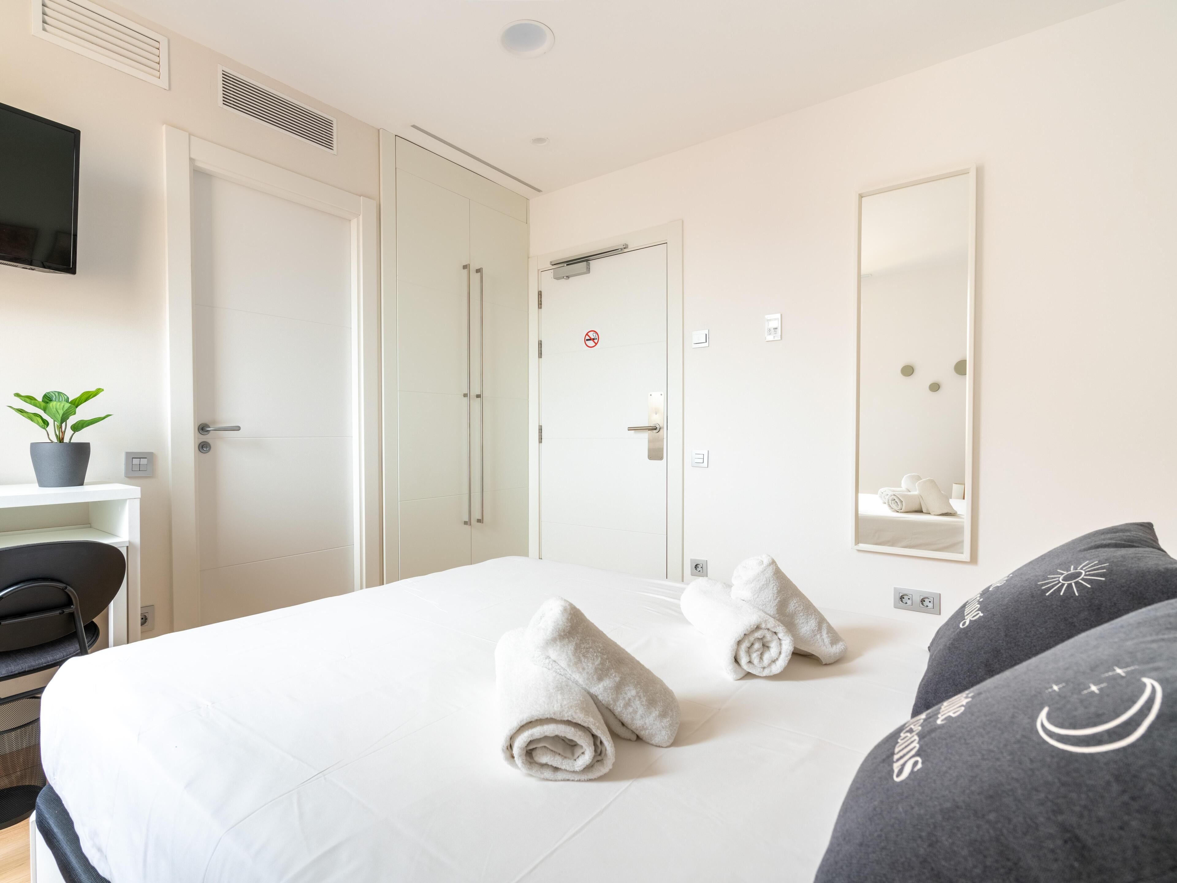 Een hotelkamer met een bed, een bureau met een stoel, een plant in pot, een spiegel en een deur met een handdoekhouder.