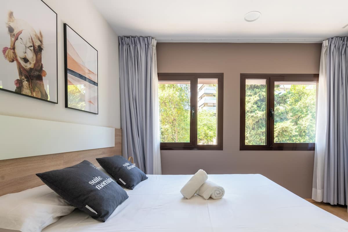 Una cama bien hecha con almohadas, una ventana con cortinas y una obra de arte en la pared.