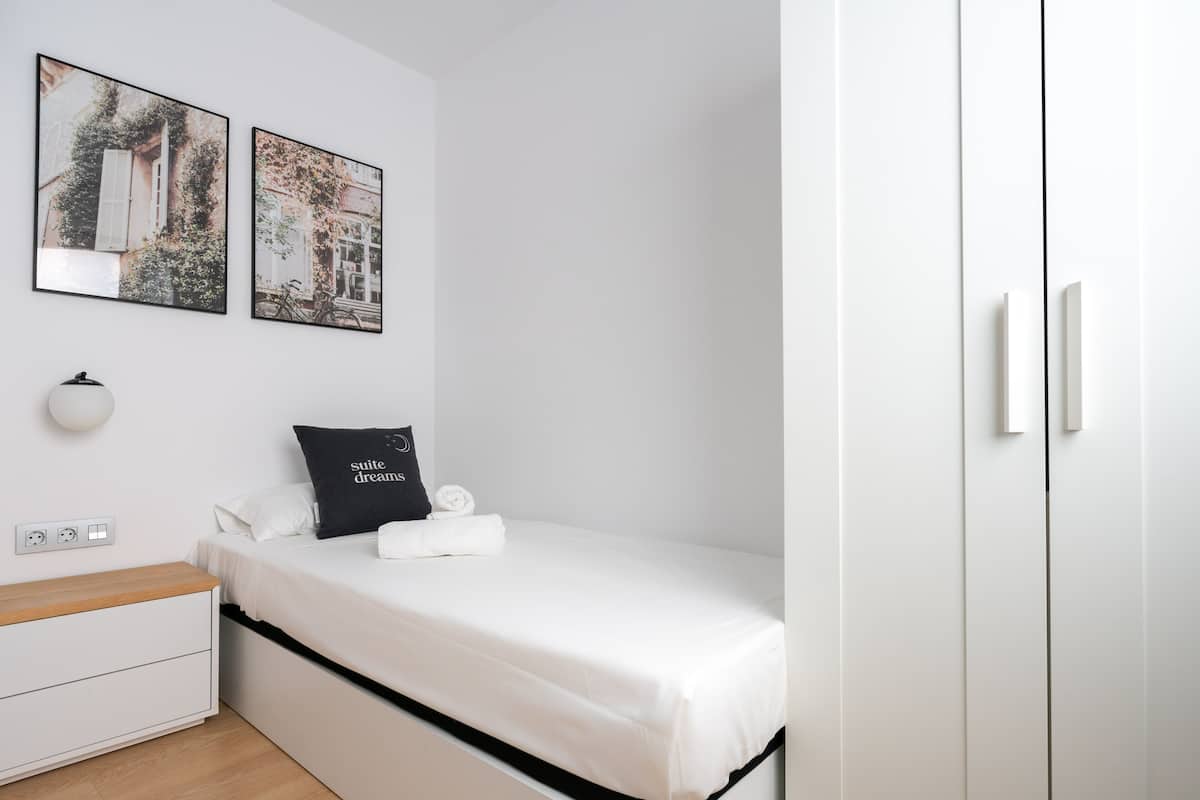 Habitación de hotel minimalista con cama, una mesita de noche, luz adosada a la pared y dos cuadros enmarcados.