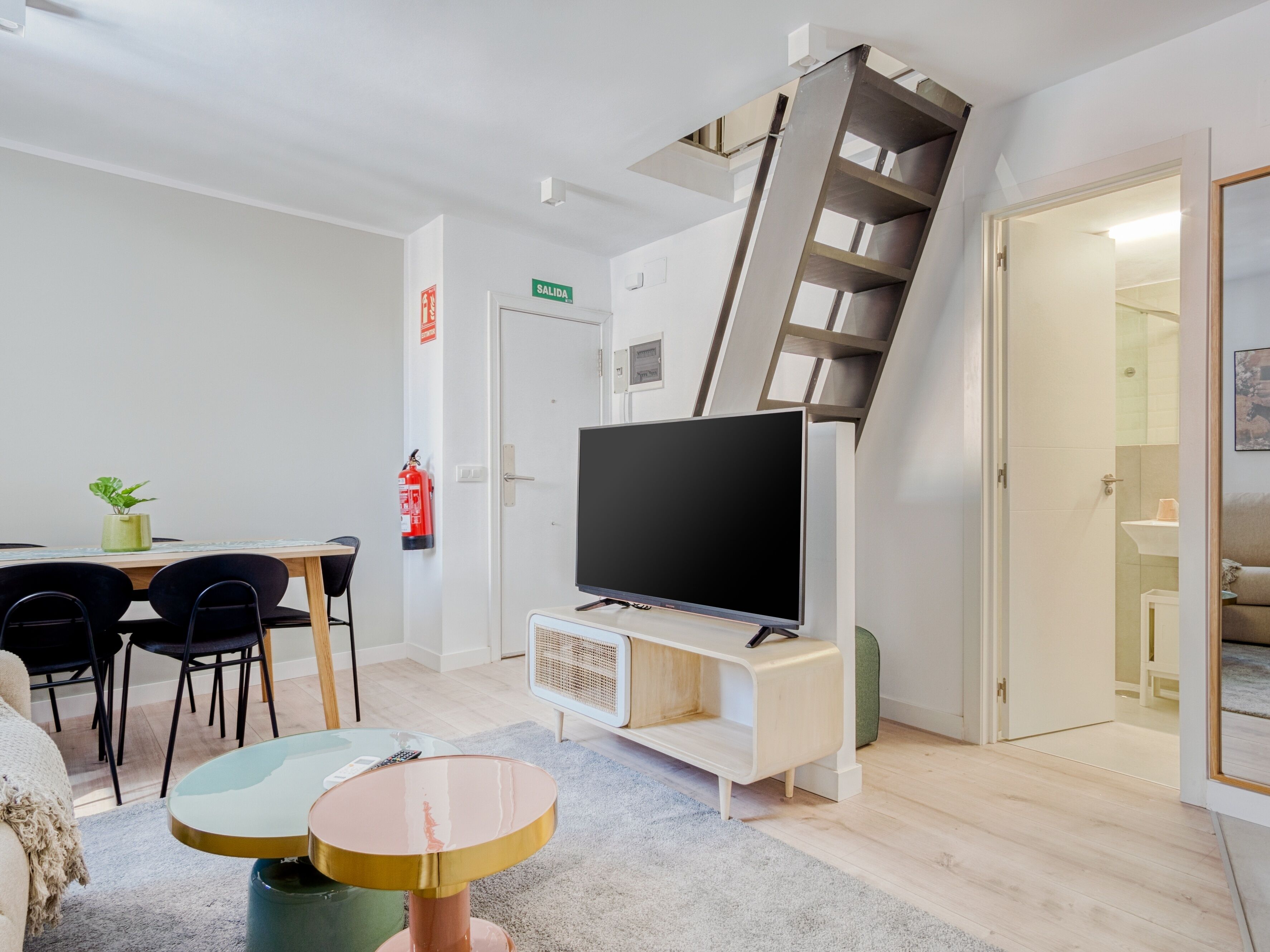 Un salon moderne doté d’un téléviseur à écran plat, d’un coin repas avec des chaises, d’un escalier et d’une porte portant l’inscription « SORTIE ».
