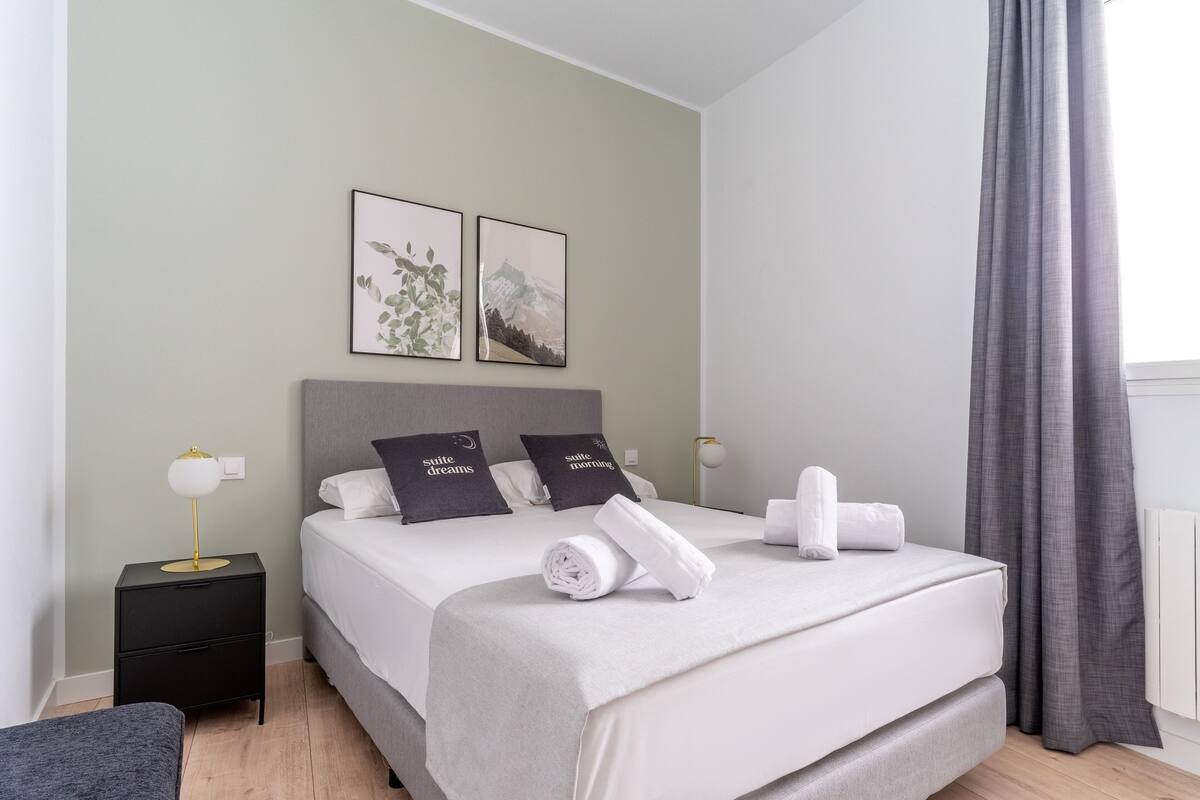 A neatly made bed with a grey headboard, two pillows with 'suite dreams' text, a small bedside table with a lamp, and a grey curtain.