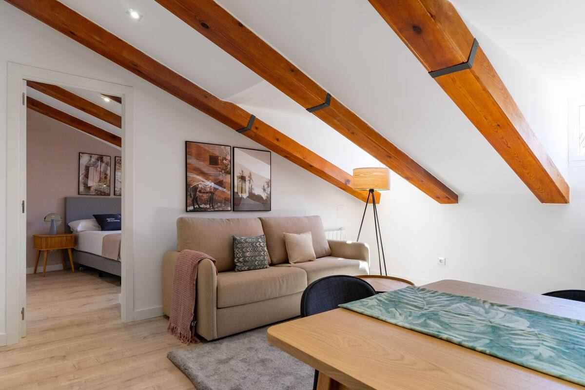 A cozy loft bedroom with exposed wooden beams, a sofa bed, a bedside table, and a floor lamp.