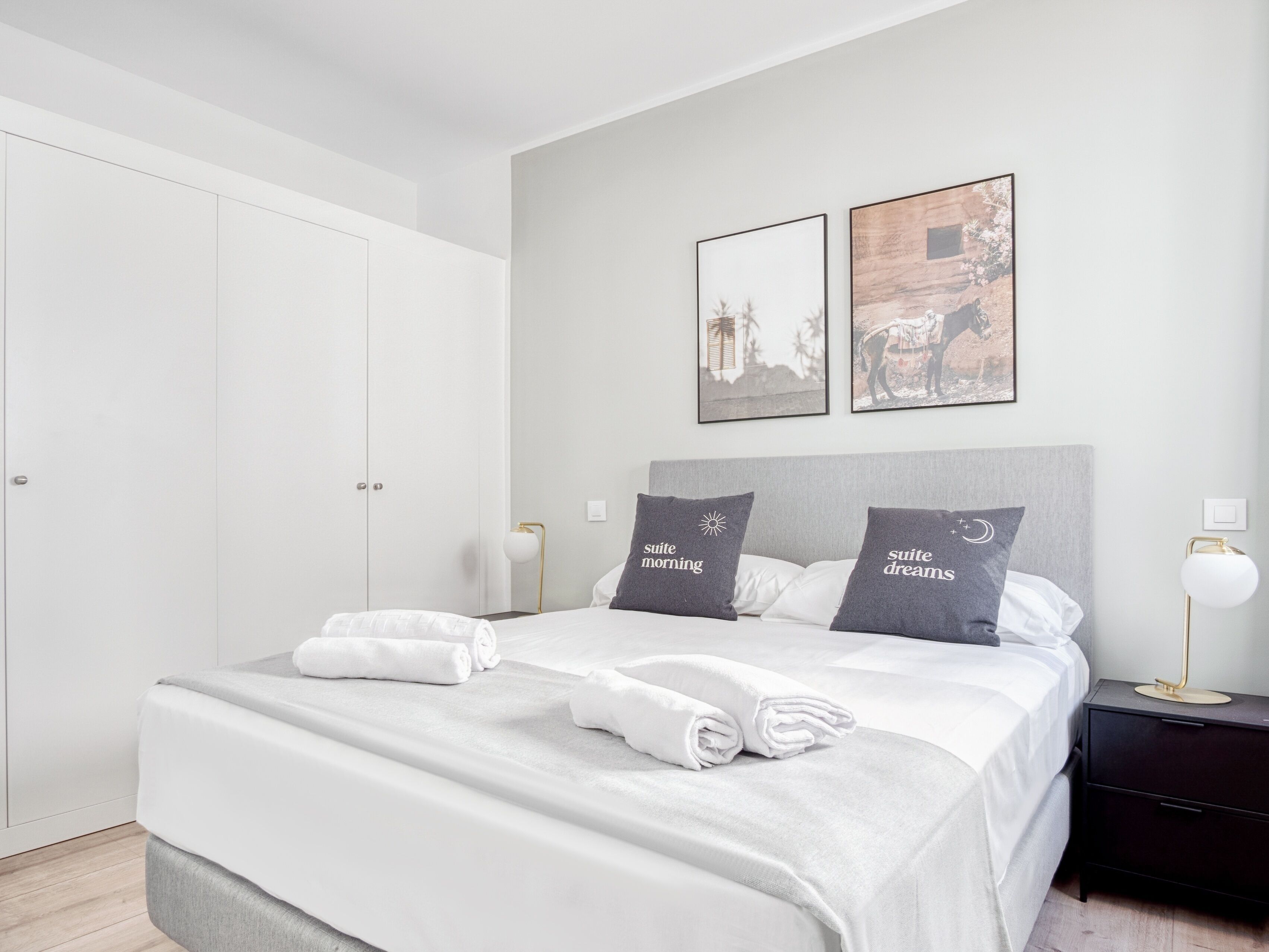 A neatly made bed with white linens and two pillows, a bedside table with a lamp, and a wardrobe.