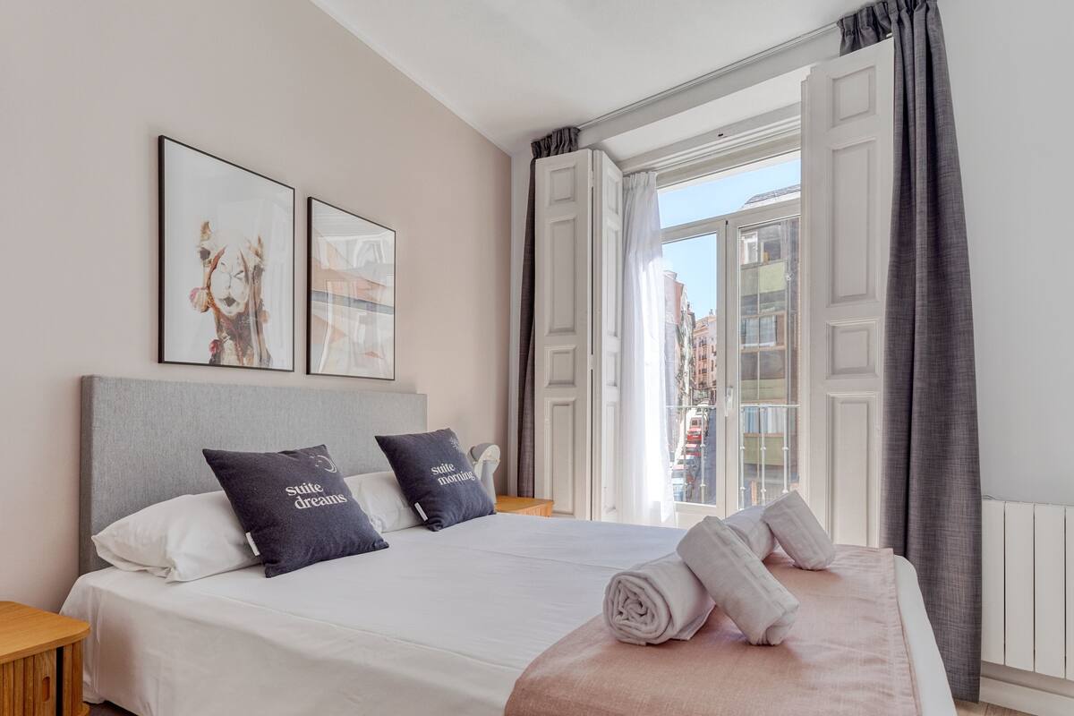 A neatly made bed with pillows, a bedside table, and a window with curtains.