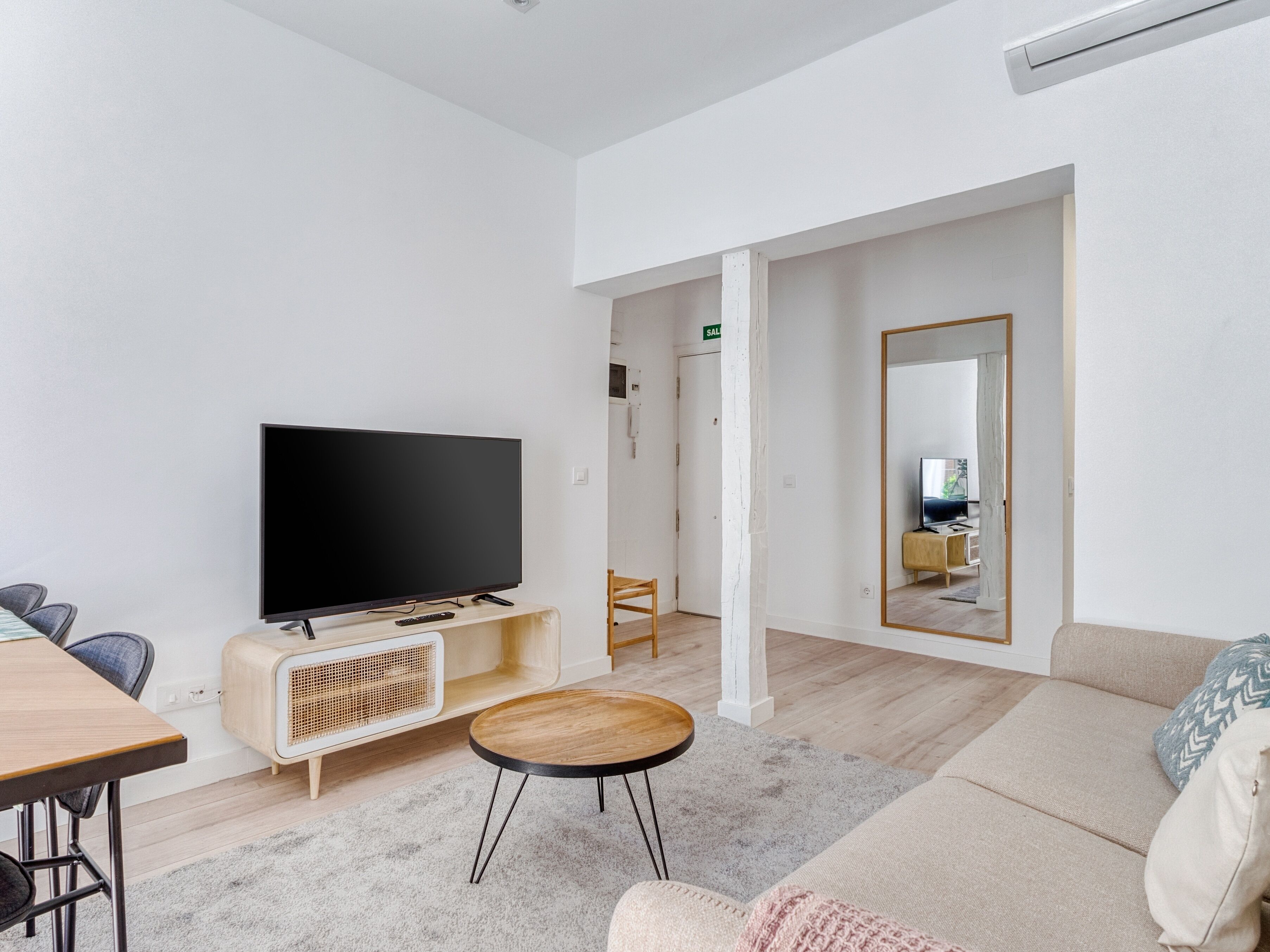Uma sala de estar moderna com uma TV de tela plana em um suporte de madeira, uma mesa de centro redonda, um sofá e um espelho grande refletindo o ambiente.