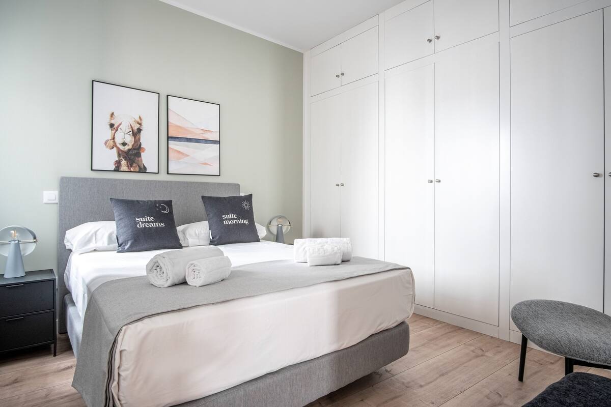 A modern bedroom with a large bed, grey and white bedding, a grey bench, and a wardrobe.