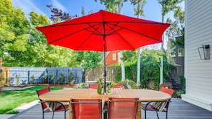 Une terrasse en bois avec une table ronde et six chaises, un parasol rouge et une maison blanche en arrière-plan.