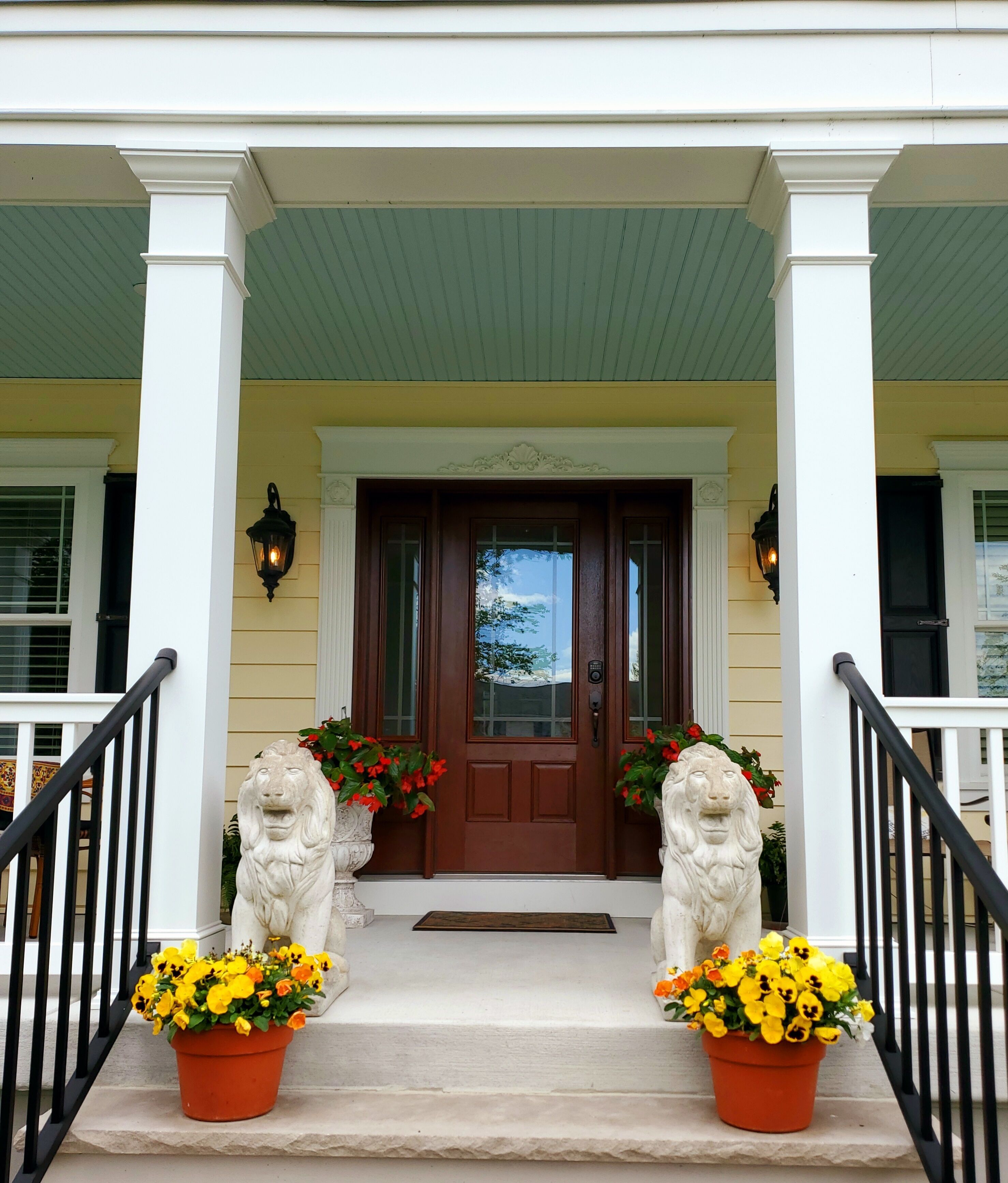 A front porch with a brown door, two lion statues, and potted yellow flowers.