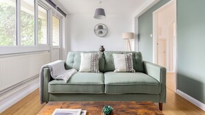 En grøn sofa med mønstrede puder, et rundt spejl, en gulvlampe og en lille potteplante på et træbord i et rum med store vinduer og en dør.