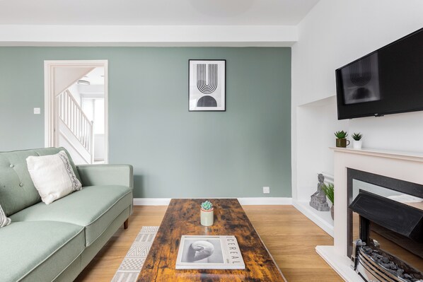 A living room with a green sofa, a wooden coffee table with a newspaper, a fireplace, and a mounted TV. -  2 Bed Flat with Garden in Canonbury (London)