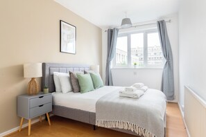 Ein ordentlich eingerichtetes Schlafzimmer mit Bett, Nachttisch, Lampe und einem Fenster mit Vorhängen.