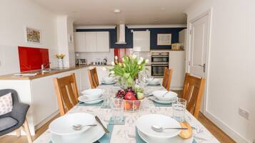 A modern dining area with a table set for four, surrounded by wooden chairs. A vase with flowers and fruits is placed in the center.