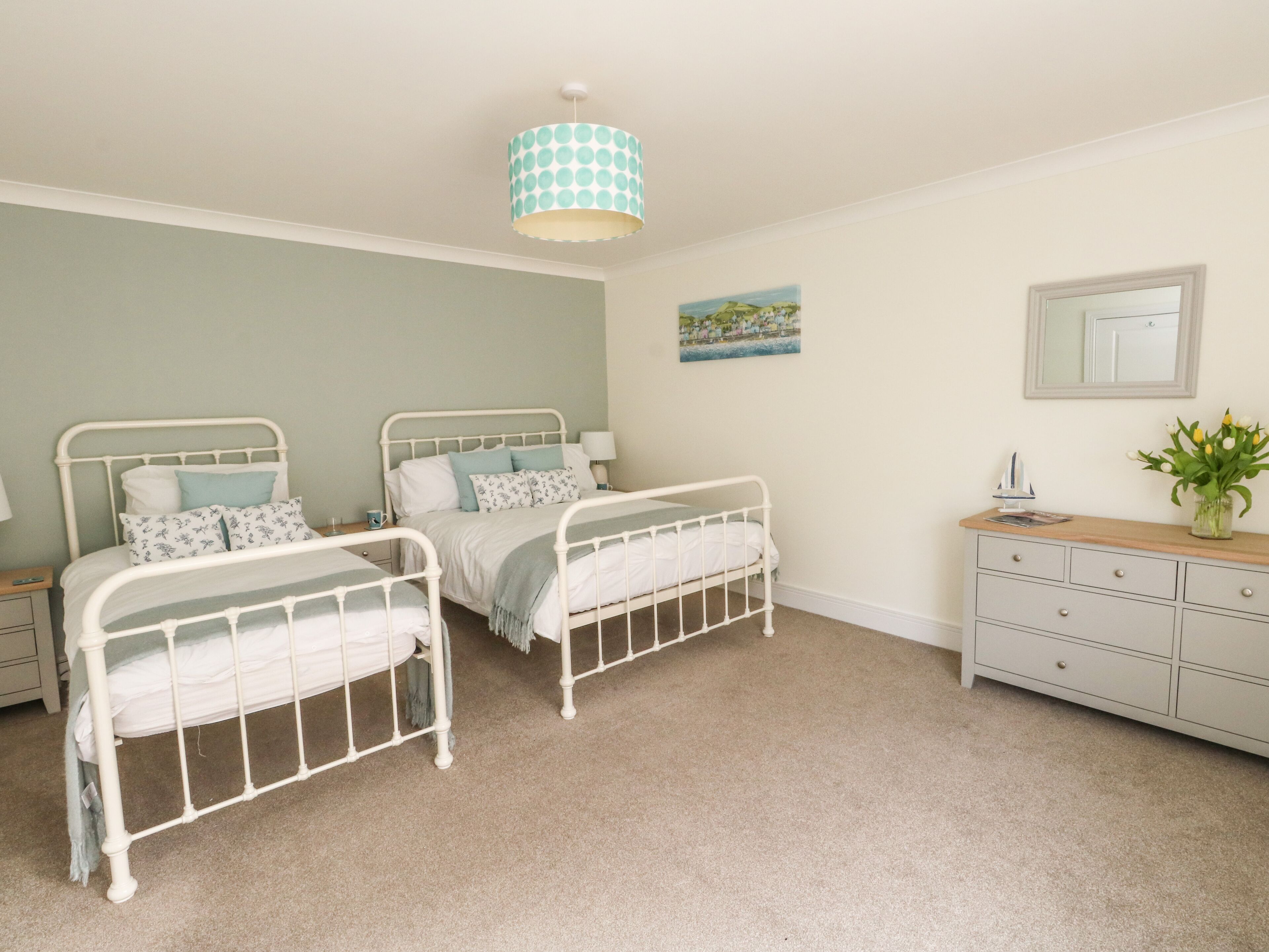 A bedroom with two white metal beds, a wooden dresser, a small table, a lamp, a painting, and a hanging light fixture.