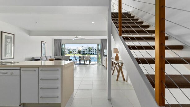 A modern kitchen with a central island, a dining area with a pool view, and a staircase with wooden railings. - Driftwood 5 Noosaville (Noosaville)