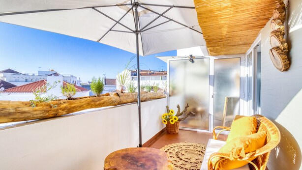 Ein Balkon mit Blick auf Gebäude, ein Korbstuhl, ein kleiner Tisch und ein Korb mit Sonnenblumen.